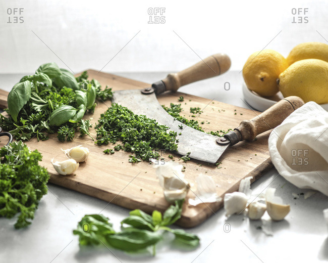 A knife with two handles resting in a pile of minced, green herbs on a wood cutting board, surrounded by lemons, cloves of garlic, and a white napkin