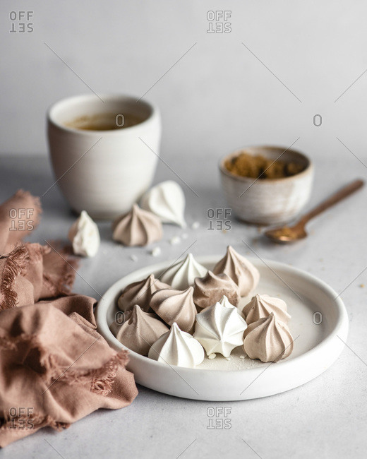 White and brown meringue cookies on a small, white dish next to a warm-colored napkin with fringed edges, with a coffee cup, a cup of brown sugar, and a rose gold spoon in the blurred background