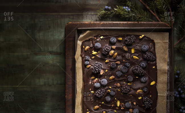 A tray of dark chocolate bark with fruit and nuts on a rustic wood board and distressed green table.
