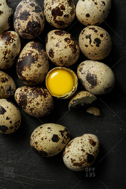 Quail eggs on a black rustic surface with one cracked open to show raw yolk inside.