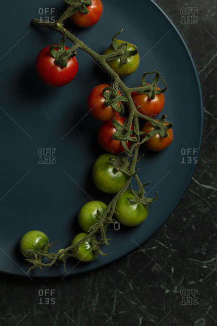 Cherry tomatoes on the vine, some ripe and some unripe, on a teal blue plate, on a green marble background.