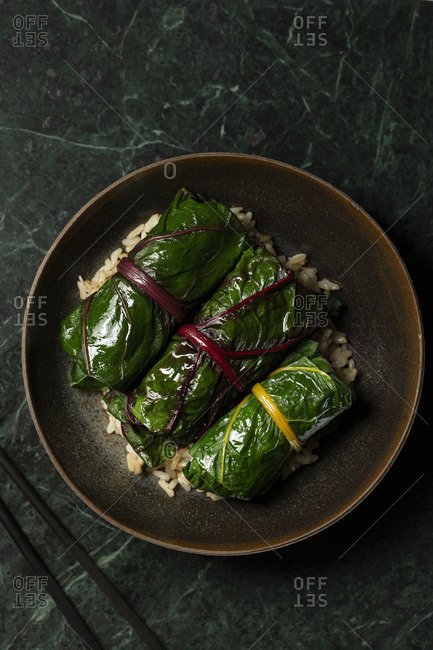 Japanese inspired rainbow chard rolls, filled with mushrooms and finished in a miso broth. They are served on  brown rice in a brown stoneware bowl, with chopsticks alongside.