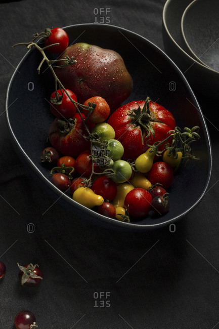 A selection of various heirloom tomatoes in a large, dark blue free form bowl. There is a stack of grey dishes in the background and loose cherry tomatoes on a charcoal linen tablecloth in the foreground.
