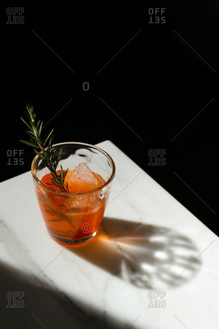Negroni cocktail with a rough ice cube and a rosemary sprig garnish on the corner of a marble table with the shadow featuring.