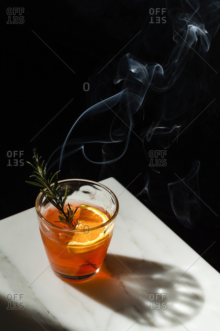 Negroni cocktail with a slice of orange and a smoking rosemary sprig garnish. The view features the corner of a marble tabletop with the shadow of the dimpled glass.