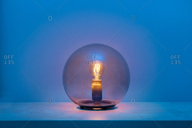Modern turned on luminaire with light bulb inside thin transparent glass sphere in middle of shelf in blue room at dusk