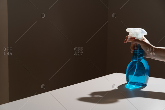 Crop anonymous person pushing dispenser mechanism and directing white cleaner flow on black wall while bottle being placed on white table with shadows