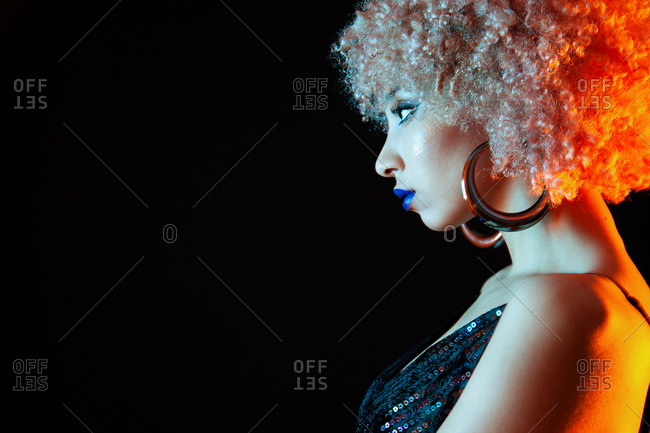 Crop stylish ethnic with blond Afro hairstyle wearing violet lipstick and elegant black dress with golden earrings looking away while standing on black background