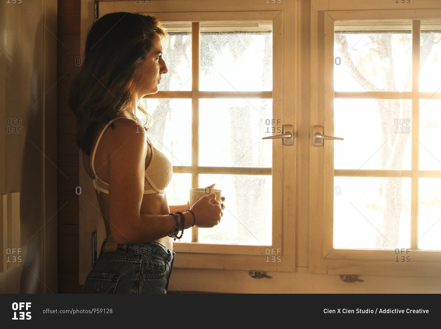 Side view of serene female in bra and jeans standing near window with cup of hot beverage and enjoying weekend in wooden cabin