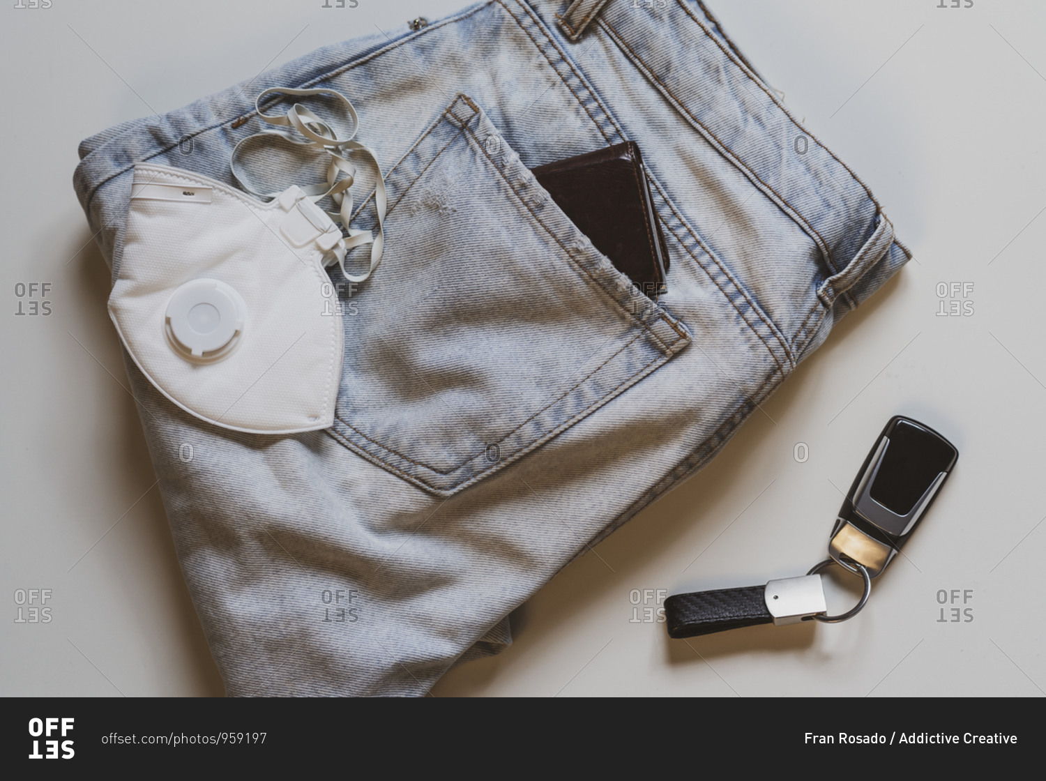From above of folded jeans with white protective mask in pocket and car keys placed on white background in light studio