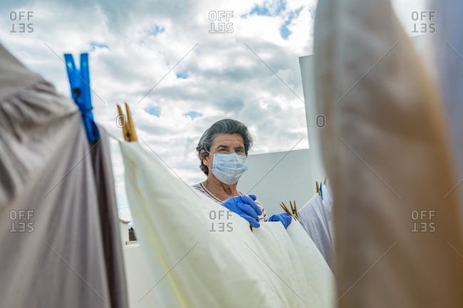 From below elderly woman looking at camera in casual clothes wearing protective mask and latex gloves hanging laundry on rope on outdoor terrace of house on cloudy day