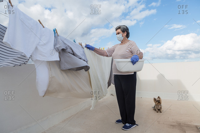 Side view of adult woman in casual clothes wearing protective mask and latex gloves holding plastic basin and taking laundry off the rope on outdoor terrace of house on cloudy day