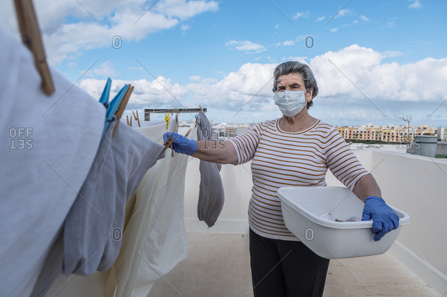 Elderly woman looking at camera in casual clothes wearing protective mask and latex gloves holding plastic basin and taking laundry off the rope on outdoor terrace of house on cloudy day