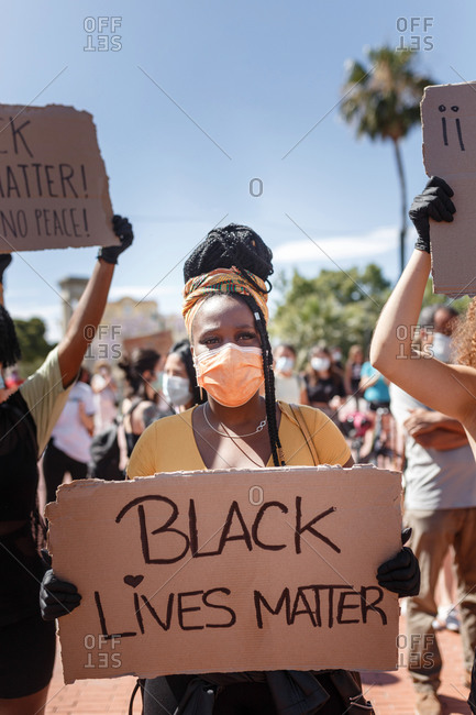 Serious ethnic female with afro hairstyle holding cardboard poster with black lives matter inscription during demonstration in crowded city and clapping