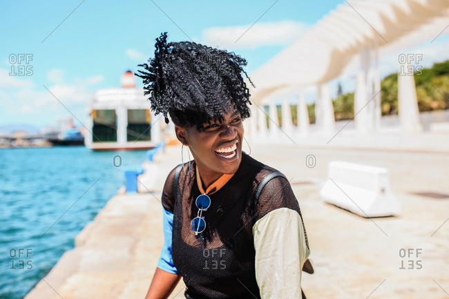 Smiling African American female with afro hairstyle standing at waterfront on sunny day and looking away