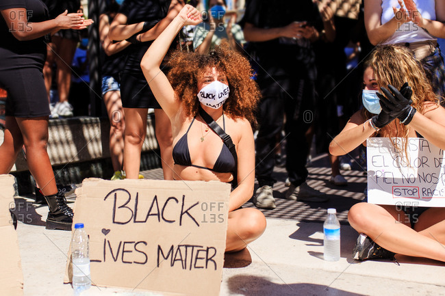 Ethnic female in medical mask sitting on sidewalk with carton poster with black lives matter inscription during protest in crowded city on sunny day