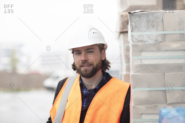 Mid adult male civil engineer leaning against stacked building material on construction site, portrait