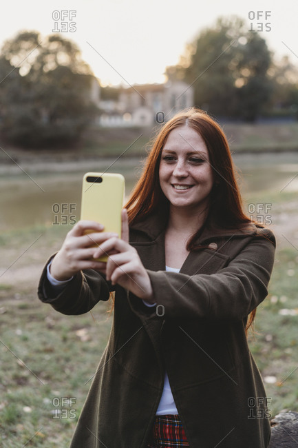 Young woman with long red hair taking smartphone selfie on riverside at dusk, Florence, Tuscany, Italy