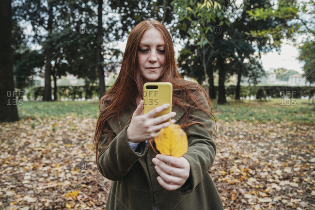 Young woman with long red hair in park taking smartphone photo of her hand holding autumn leaf