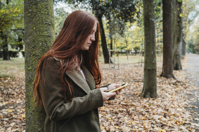 Young woman with long red hair in autumn park looking at smartphone