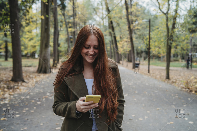Young woman with long red hair looking at smartphone in tree lined autumn park, Florence, Tuscany, Italy