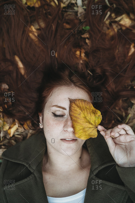 Young woman with long red hair lying amongst autumn leaves and covering eye with autumn leaf, overhead portrait
