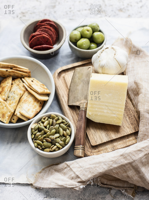Still life of cutting board and cheese with bowls of crackers, olives, salami and pumpkin seeds, overhead view