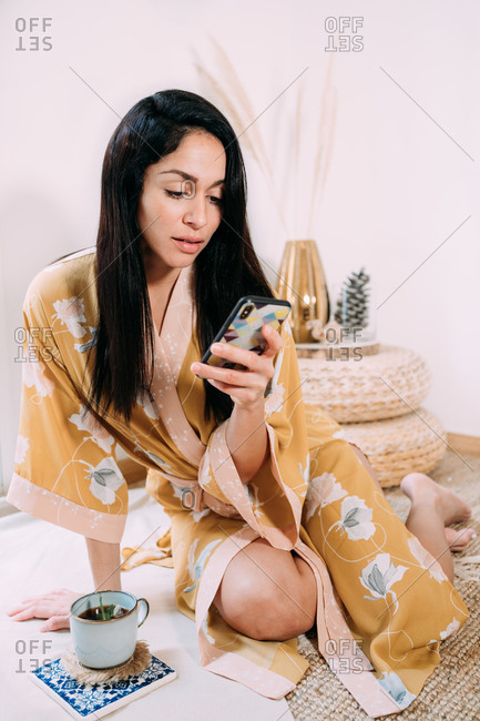 Gorgeous young lady in sleepwear sitting on floor with cup of aromatic tea and texting messages on mobile phone while spending weekend at home