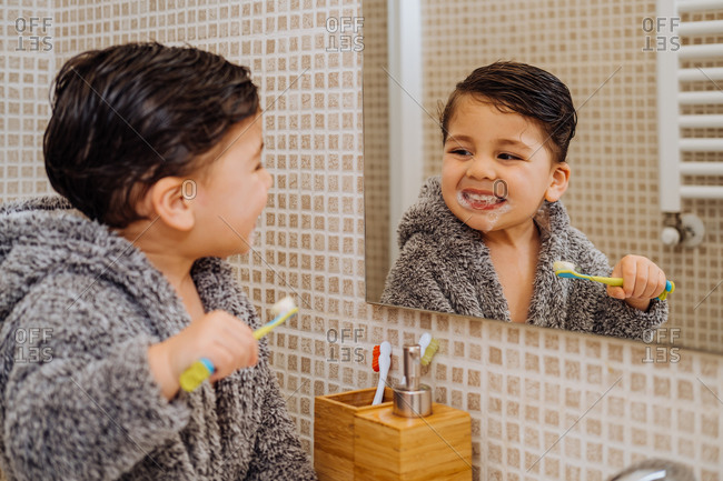 Adorable child wearing cozy bathrobe standing in bathroom with toothbrush and looking in mirror