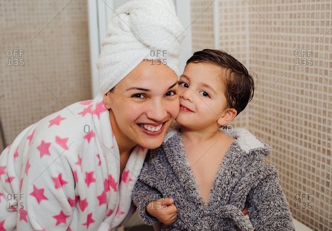 Cheerful woman with towel turban cuddling little child in bathrobe after taking shower and looking at camera