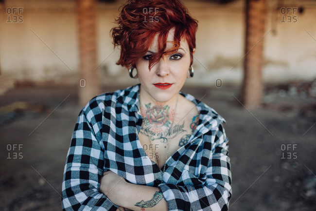 Independent stylish young female with trendy haircut and tattoos wearing casual checkered shirt looking at camera while standing against blurred shabby building