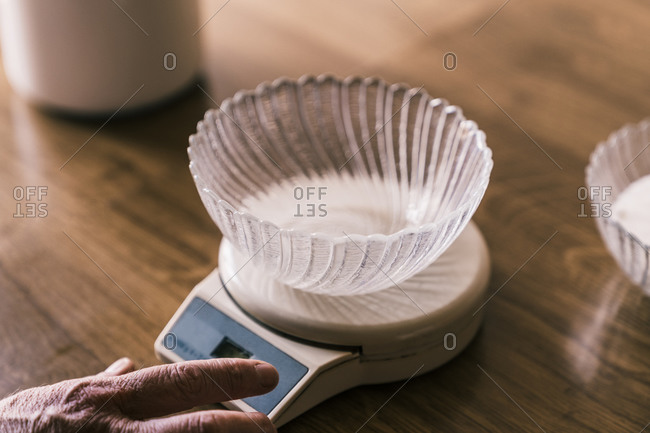 Cropped unrecognizable female hand weighting with electronic kitchen scale with glass bowl on top on wooden table