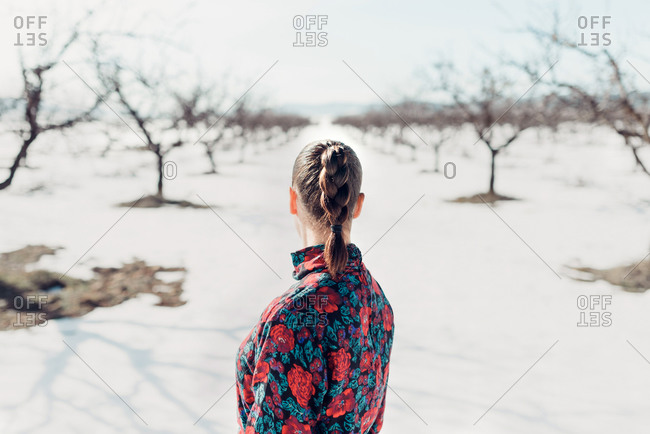 Back view brunette female in stylish colorful outfit while standing in snowy field with leafless trees in sunny winter day