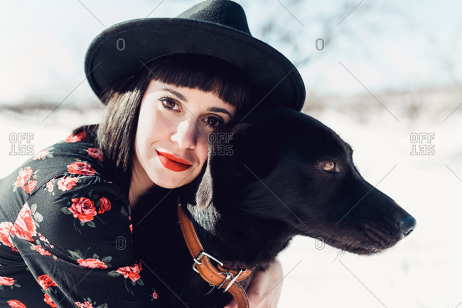 Smiling young fashionable brunette female in black hat and dress and beautiful black dog looking at camera in snowy field with leafless trees in sunny winter day