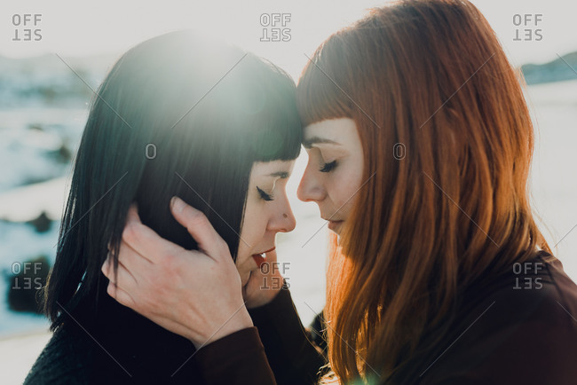 Side view of modern sensual lesbian couple in love embracing while standing in back lit in snowy field at countryside