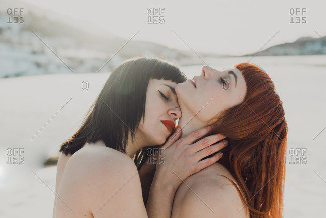 Side view of sensual tender young women in love with bare shoulders embracing and kissing while standing in back lit in snowy field