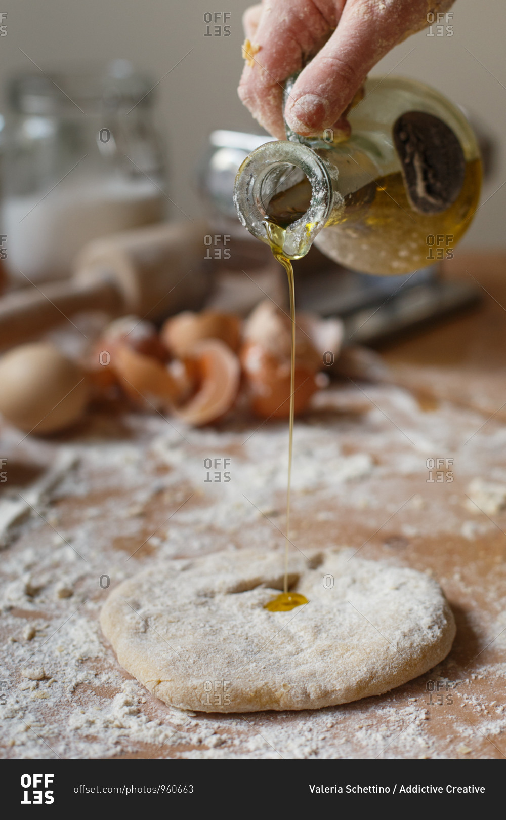 Faceless cook pouring fragrant fresh olive oil from glass jug onto rolled pasta dough on background of blurred kitchen mess