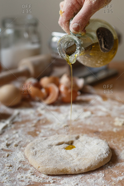 Faceless cook pouring fragrant fresh olive oil from glass jug onto rolled pasta dough on background of blurred kitchen mess