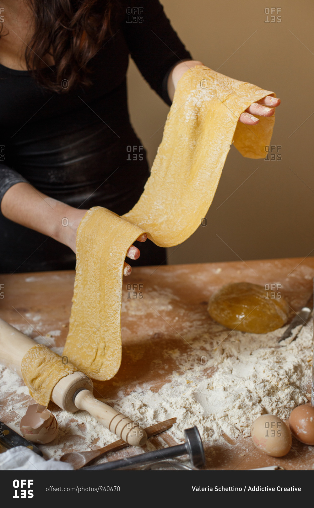 Anonymous lady in black dress holding finely rolled pasta dough in hands and standing near messy table while cooking Italian dish at home