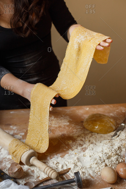 Anonymous lady in black dress holding finely rolled pasta dough in hands and standing near messy table while cooking Italian dish at home