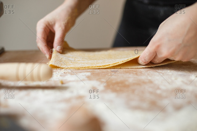 Unrecognizable cook in black wear folding rolled thin pasta dough into several layers during preparation of pastry on table with flour