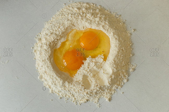 Top view of flour pile with broken raw egg in middle on wooden kitchen table with pasta ingredients and kitchen utensil on bright sunny day