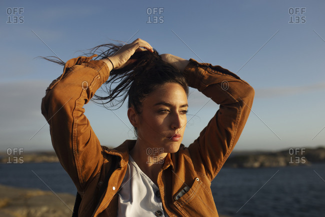Beautiful young female traveler in casual clothes standing on rocky seashore holding hair up with hand in a high ponytail during a windy day and looking away dreamily