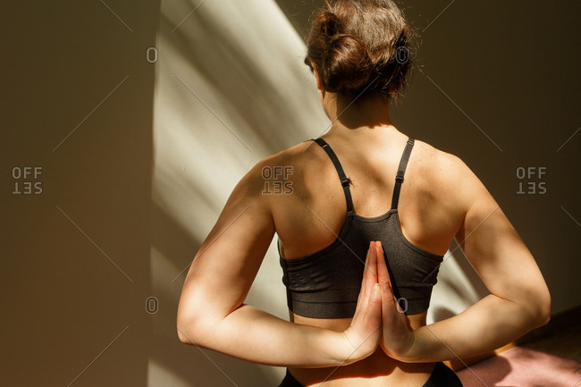 Crop back view of unrecognizable female in black sports bra practicing behind namaste yoga pose at home in sunlight