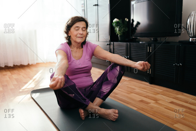 Focused senior woman doing yoga at home. 70 years old woman sitting on  fitness mat with outstretched hands resting on her knees performing yoga in  living room. stock photo - OFFSET
