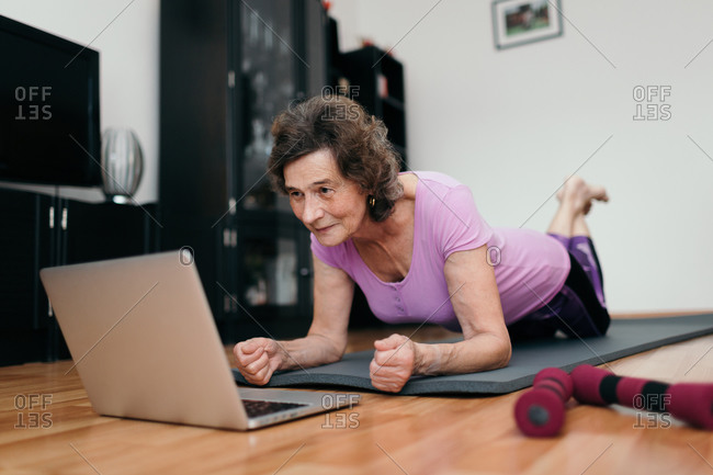 Elderly woman exercising at home watching online course. Low angle shot of senior woman at her 70s lying on floor exercising plank while watching fitness training online on notebook at home.