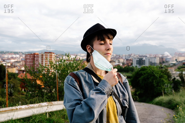 Serious young lady in casual clothes and hat standing with medical mask on street with eyes closed and enjoying fresh air during walk being on quarantine