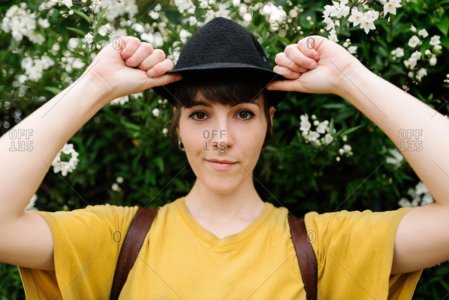 Positive young stylish lady in casual yellow shirt and stylish black hat standing near blooming bushes and looking at camera