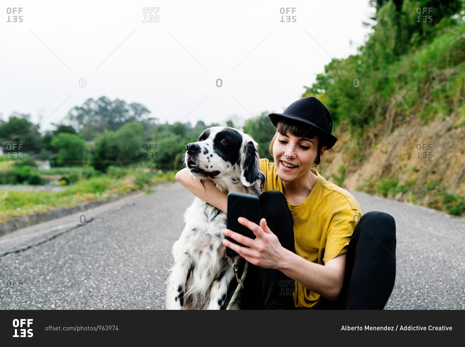 Cheerful young lady in casual clothes and hat sitting on ground with mobile phone and taking selfie with dog during walk on street
