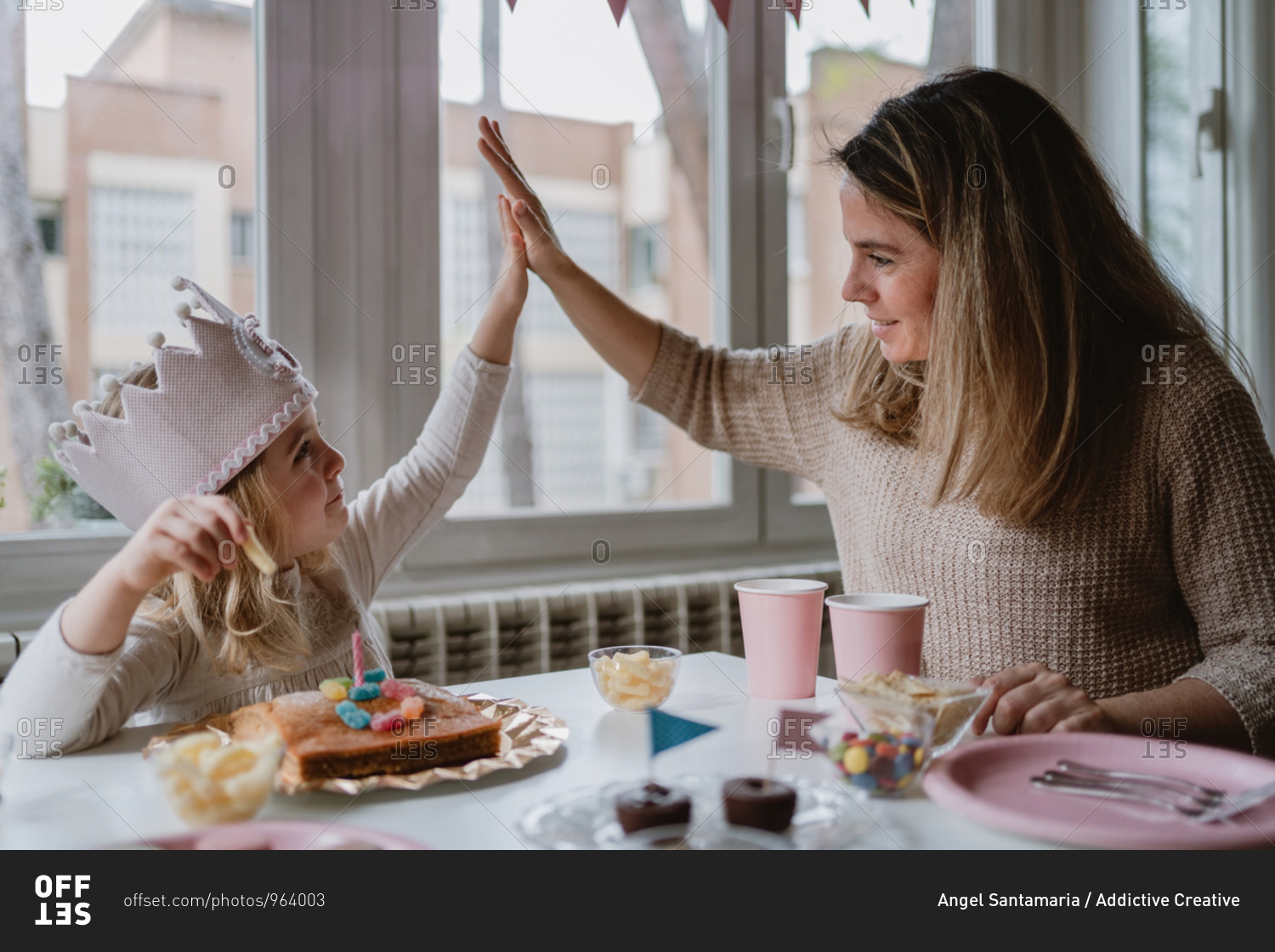 Cute little girl in casual clothes and paper crown giving high five with mother while sitting at table and eating delicious food during holiday